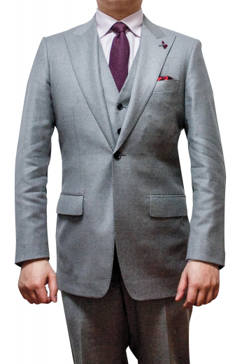 Mens made to order light grey 3pc suit in cashmere wool. Features a bespoke slim fit vest with 6 front buttons, a handmade jacket with 2 peak lapels and 1 front close buttons, and tailor made slim fit dress pants with sewn down pleats. The mens handmade slim fit jacket has 1 boutonniere on the left lapel, 1 upper welt pocket, 2 lower flapped pockets, and hand moulded shoulders. The mens bespoke vest has 2 welted lower pockets. Mens handmade dress pants have 2 front slash pockets, a 2 point button and hook closure, and a zip fly.